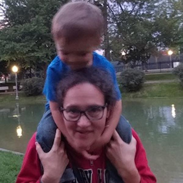 Elena Nicoladis with a child on her shoulders. She is holding the childs knees and wearing a red shirt and glasses, and standing in front of a pond with lights in the background