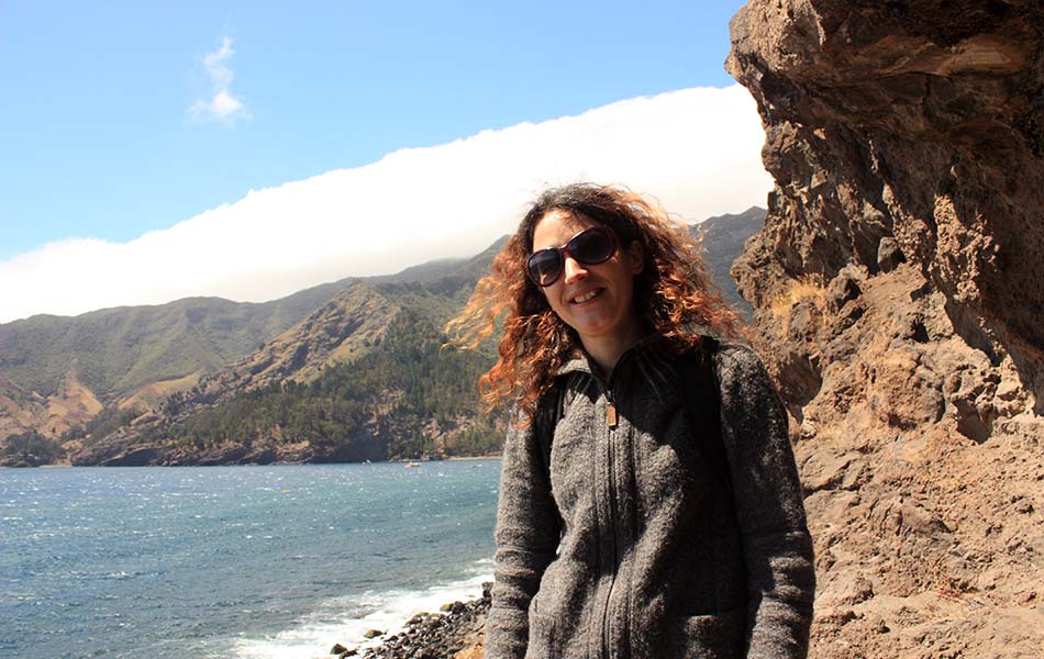A photo of Dr. Dianne Newbury standing on the shores of Robinson Crusoe Island, with the ocean and part of the island in the background. She is wearing a grey jumper and sunglasses.