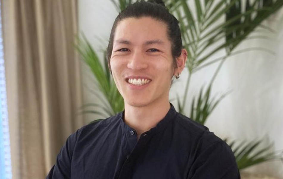 A headshot of Andrew Cheng standing with arms crossed, smiling at the camera, wearing a black shirt. Behind is palm plant and beige curtains