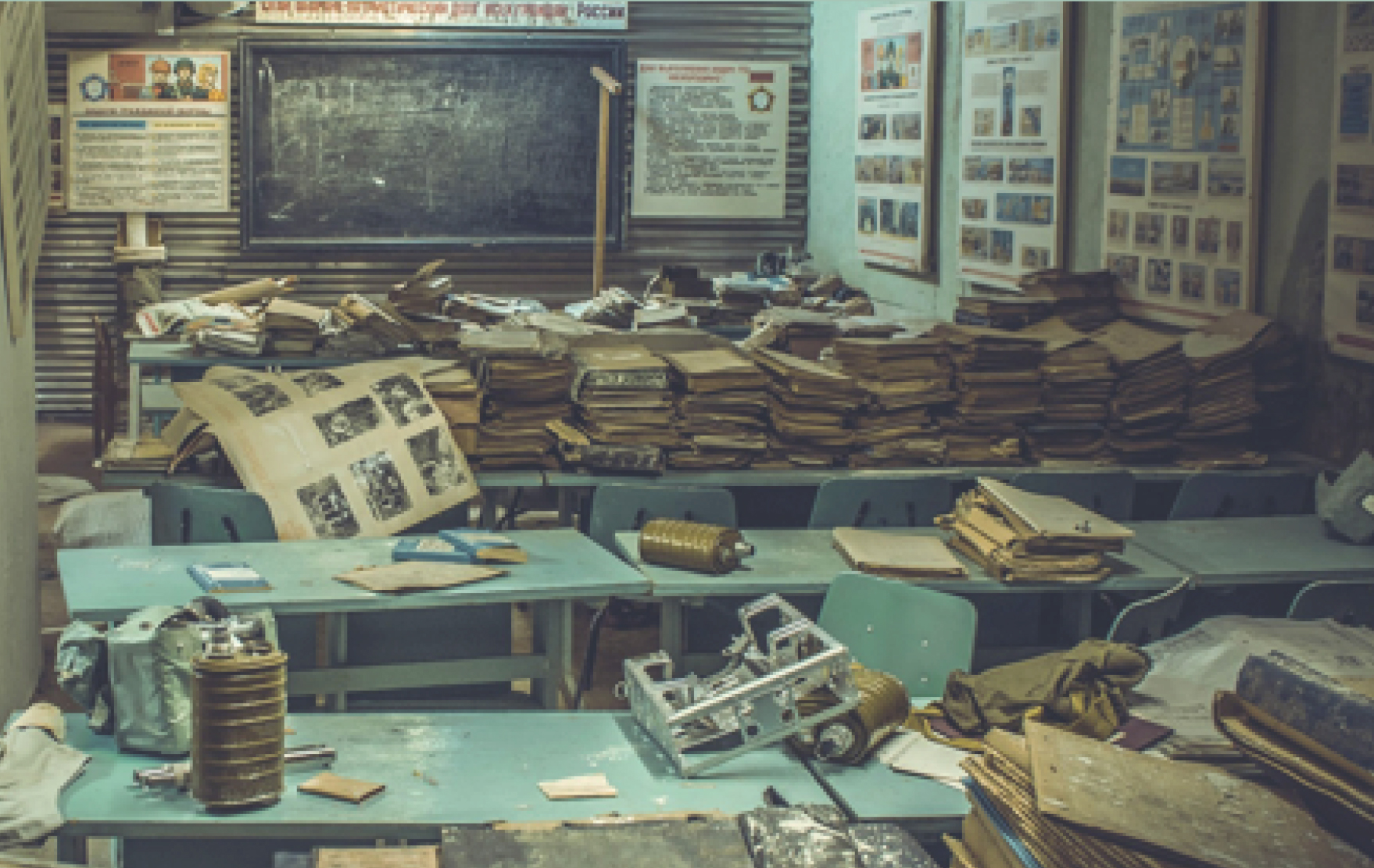 A picture of a cluttered classroom with a blackboard at the front, green tables and desks piled with books and papers, and posters on the walls.