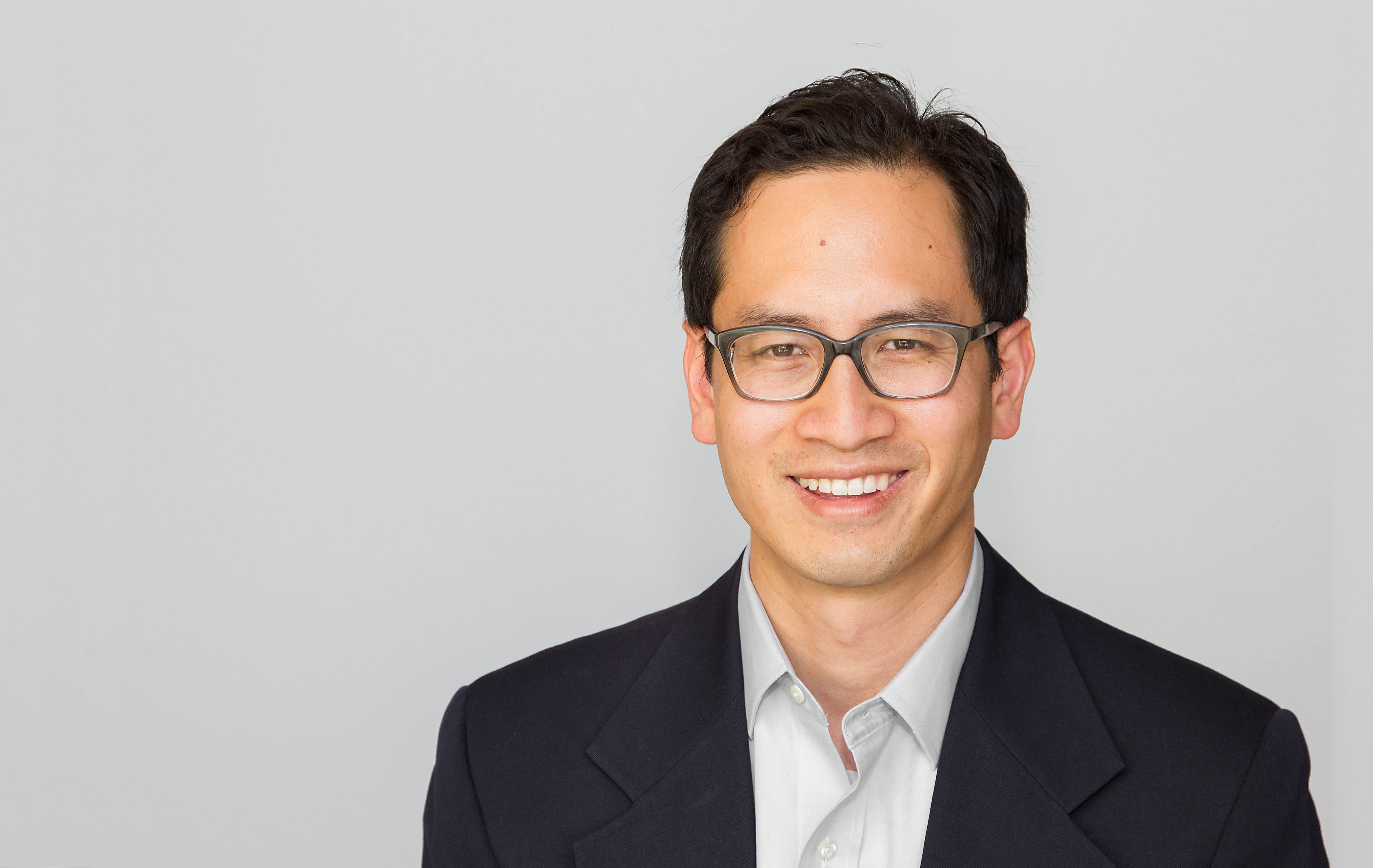 A headshot of Professor Edward Chang, smiling, wearing glasses, and standing against a grey background.