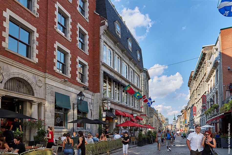 An image of a downtown Montreal street lined with flags, with a brick building on the left, people sitting on the street at tables, and people walking down the street