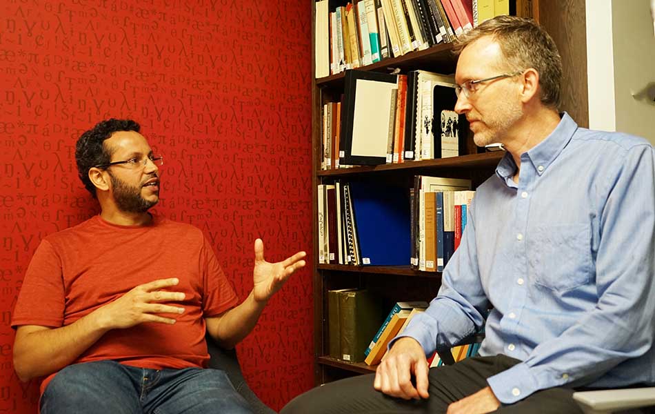 A picture of assistant professor Muhammad Abdul-Mageed speaking and Professor Bryan Gick listening against a red wall, with a bookcase in the background