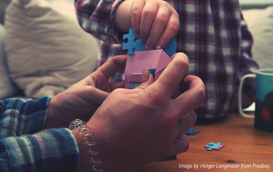 A close up on an adult's hands holding a pink box, with a child's hand placing a blue puzzle piece in the box