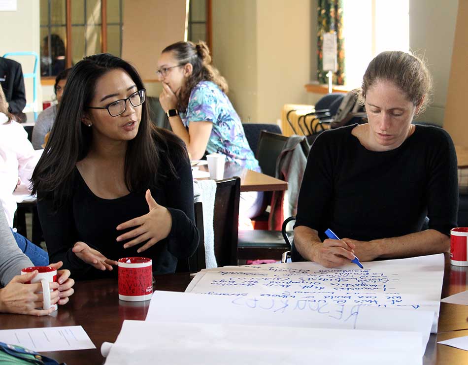 Two language sciences members sit at a table, discussing research at a recent member event.  One of them, a woman with long dark hair, glasses, and a dark-colored long-sleeved shirt, gestures while speaking, while the other, a woman with light-coloured hair pulled back in a ponytail, wearing a dark-coloured short-sleeved shirt,  writes down notes on a large flipchart on the table in front of them.  Other language sciences members can be seen conversing in the background.