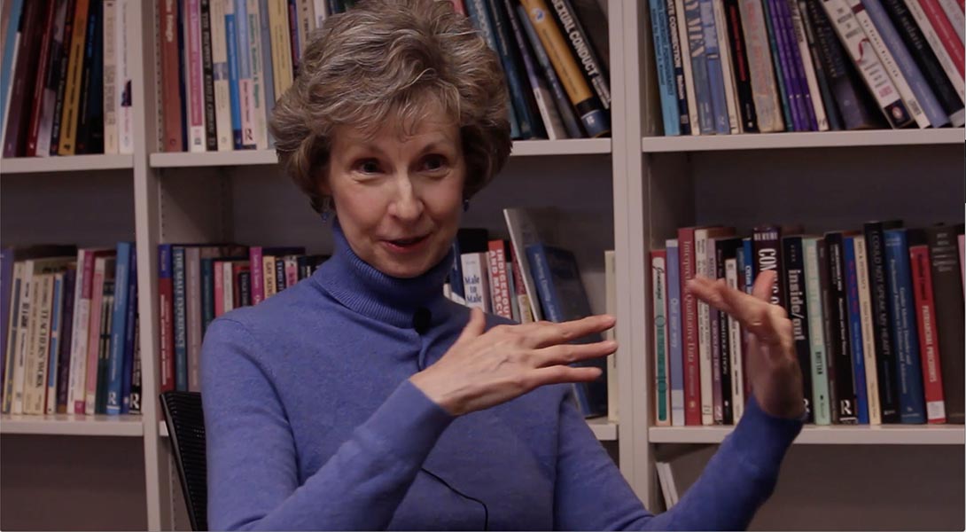 A picture of Professor Janet Jamieson wearing a purple sweater sitting in front of a bookcase, midway through the ASL sign for Star Wars