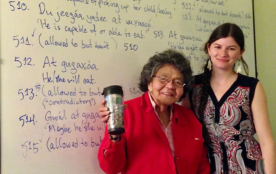 A picture of Elder Keiyishí Bessie Cooley, wearing a red jumper and raising a coffee travel mug, and Heather Burge wearing a dress with her arm around Elder Cooley. Both are standing in front of a whiteboard with Lingít and English on it