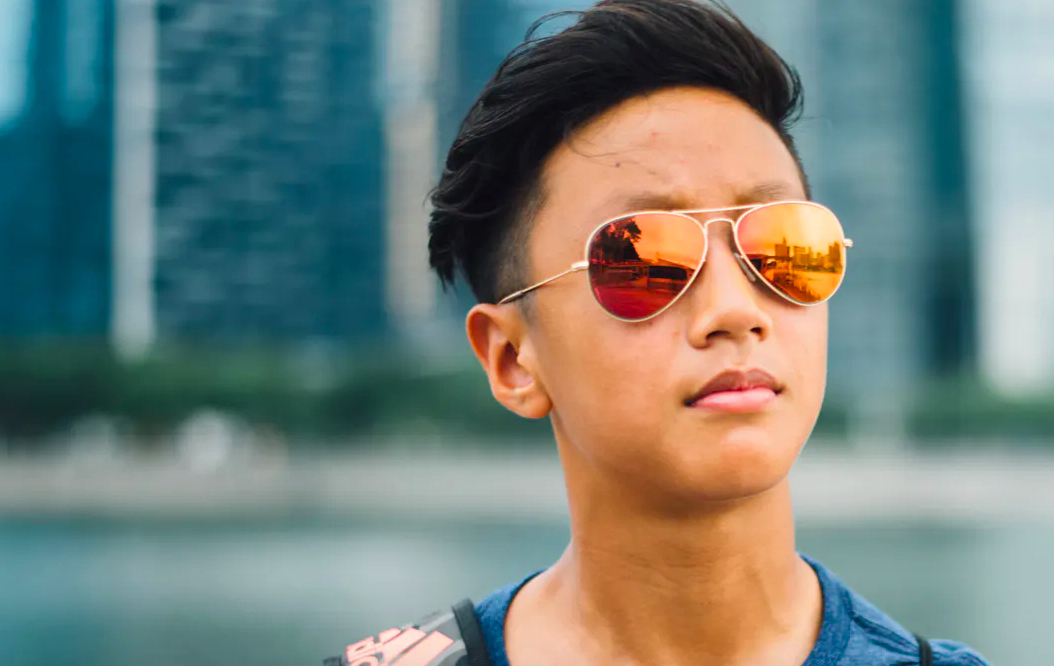 A picture of a young person of Asian heritage wearing sunglasses and standing in front of a harbour