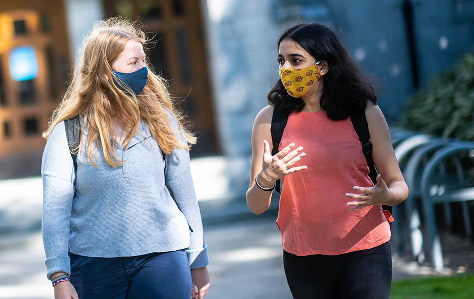 An image of two students walking on UBC campus, wearing masks and with long hair. One student is gesturing as they talk.