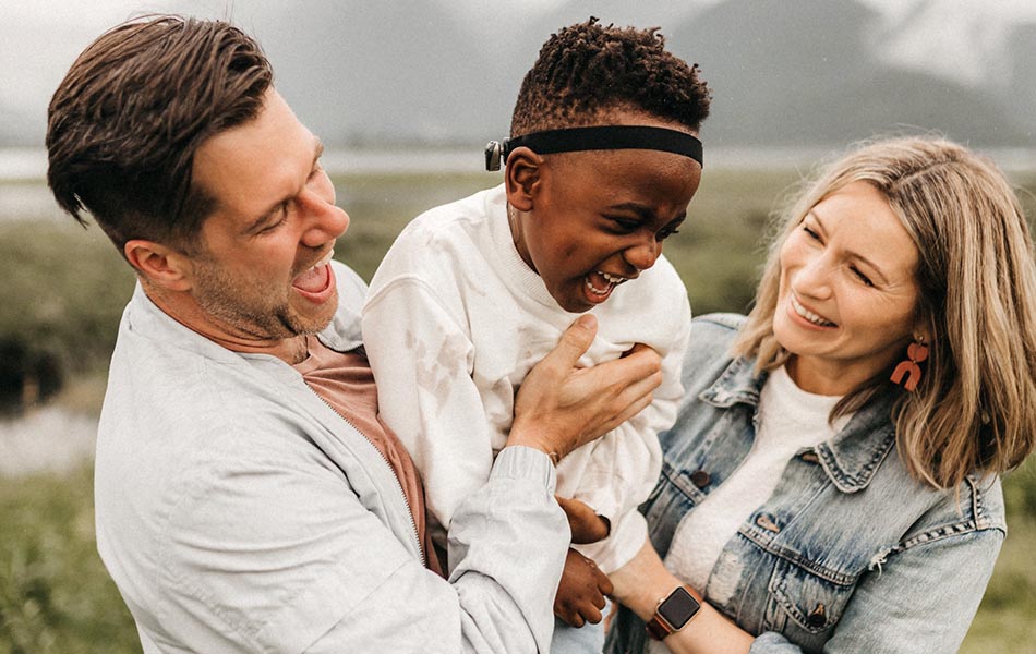 A picture of a mother and father lifting their child up in front of mountains. Everyone is laughing.