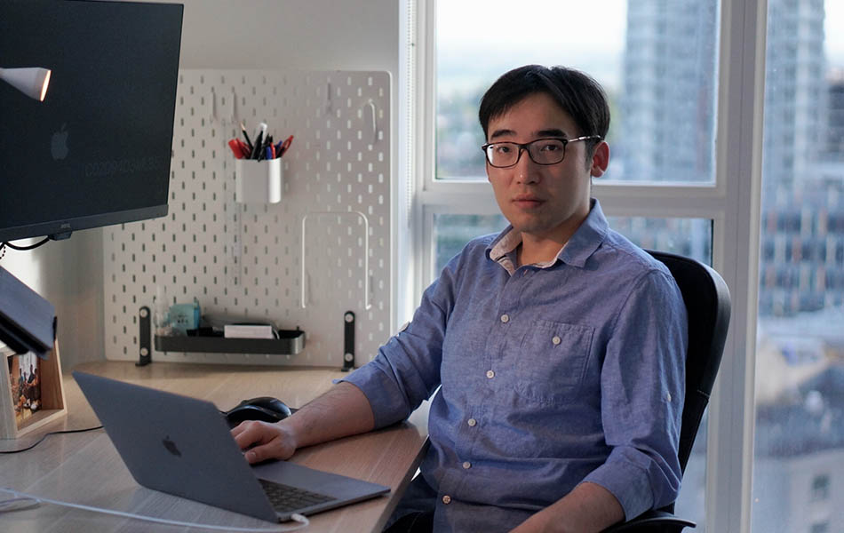 Keyi Tang sitting at a desk in front of a latop and a window with a cityscape. He is wearing a blue shirt and glasses