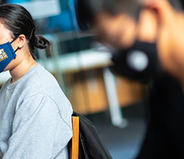 An image of students wearing masks, working at computers. One is wearing a blue mask with the UBC logo and a grey sweater. The other is wearing a black sweater.