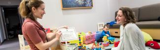 A picture of postdoctoral fellow Dr Rebecca Reh presenting a baby scientist certificate to a mother, while her child plays with toys in the background