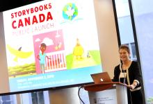 An image of Bonny Norton standing at a podium in front of a projector screen, at the launch of Storybooks Canada, 
