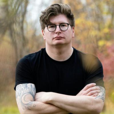 A head shot of Dallas Hunt standing with arms crossed in front of fall foliage, wearing a black shirt and glasses
