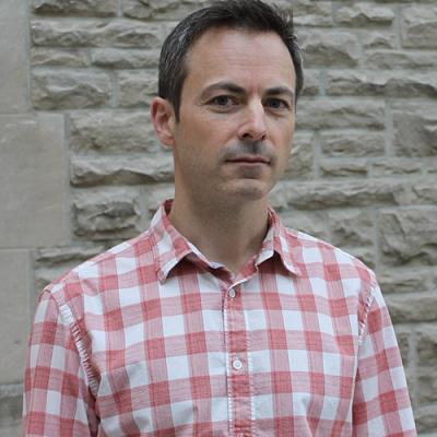 A headshot of Marc Joanisse standing in a checked shirt against a sandy coloured brick wall.