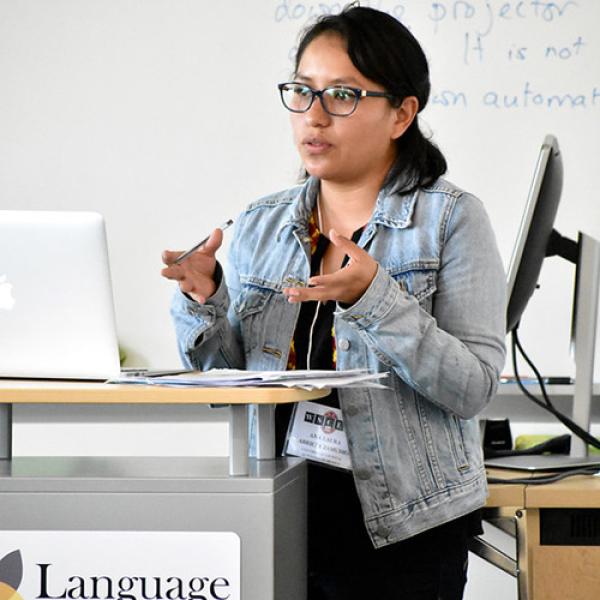 A picture of Ana Laura Arrieta Zamudio standing at a podium, gesturing with her hands. She is standing in front of a white board and laptop is on the podium. She is wearing a denim jacket and glasses.