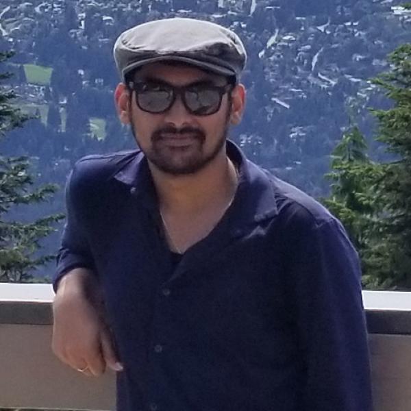 A picture of Venkata Praneeth Srungarapu wearing sunglasses and a hat, standing against a wooden fence with a view over Vancouver harbour in the background.